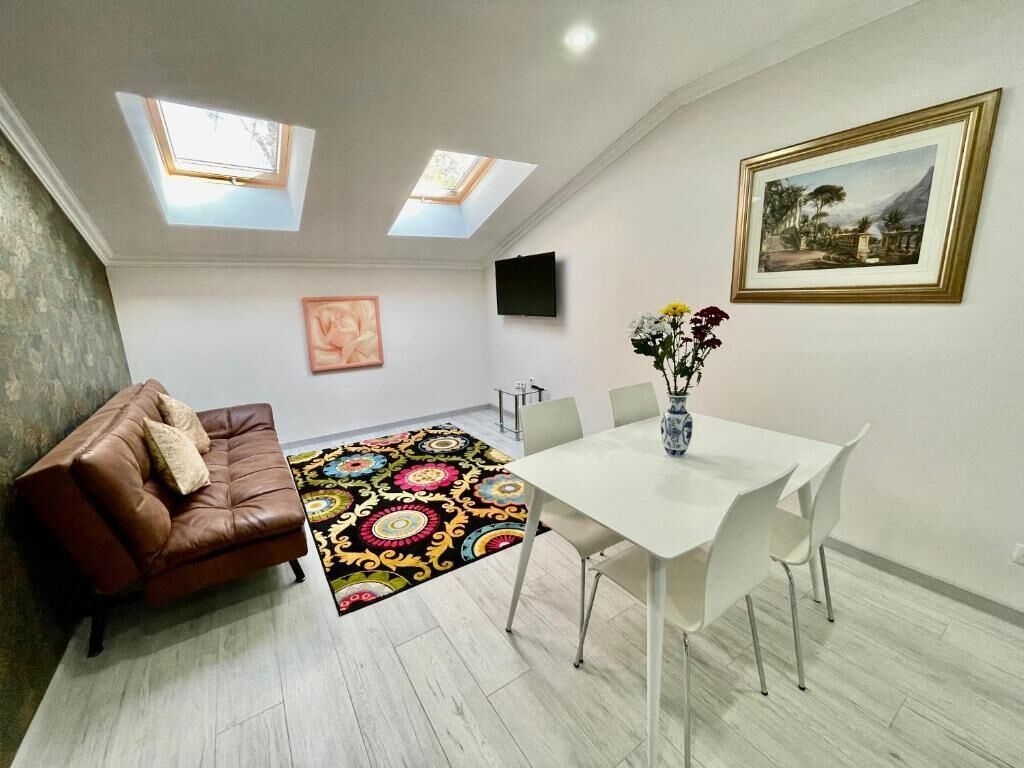 Een kamer met een bruine leren bank, een witte eettafel met stoelen, een kleurrijk vloerkleed en een schilderij in een lijst aan de muur.