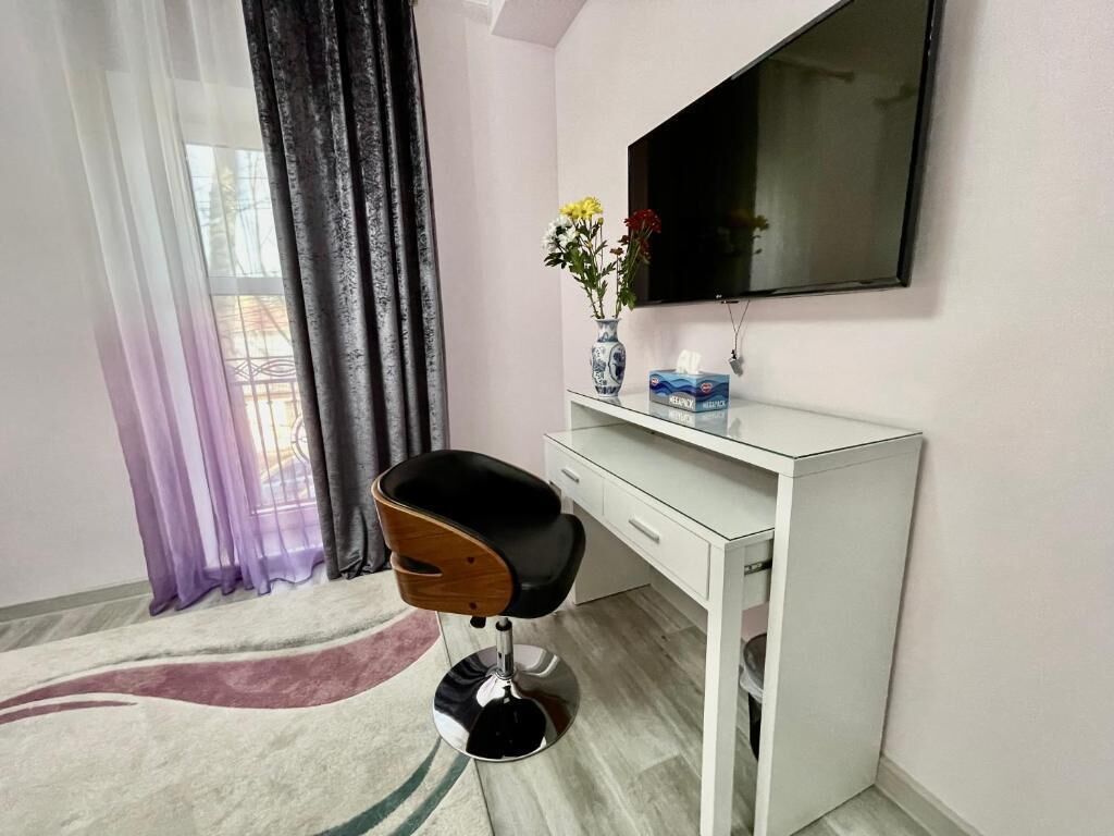 Una habitación moderna con escritorio blanco, silla negra, televisor de pantalla plana montado en la pared, un jarrón con flores y una ventana con cortinas translúcidas.