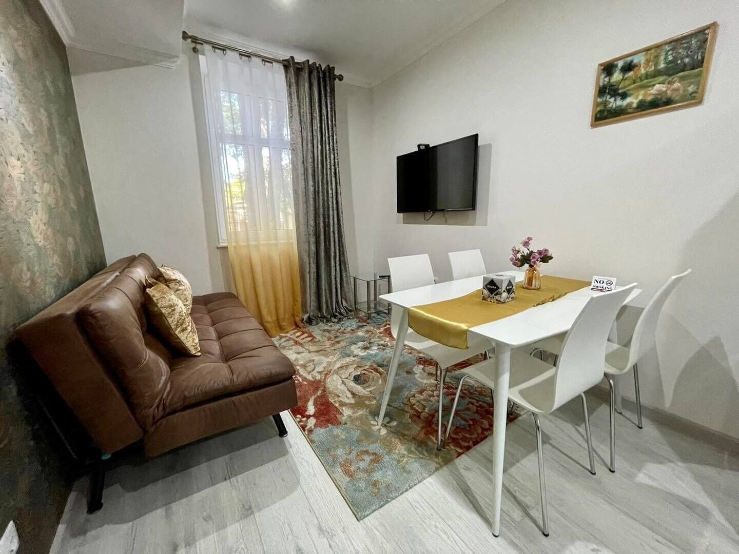 Een moderne woonkamer met een bruine leren bank, een witte eettafel met vier stoelen, een flatscreen televisie aan de muur en een vloerkleed met bloemenmotief.