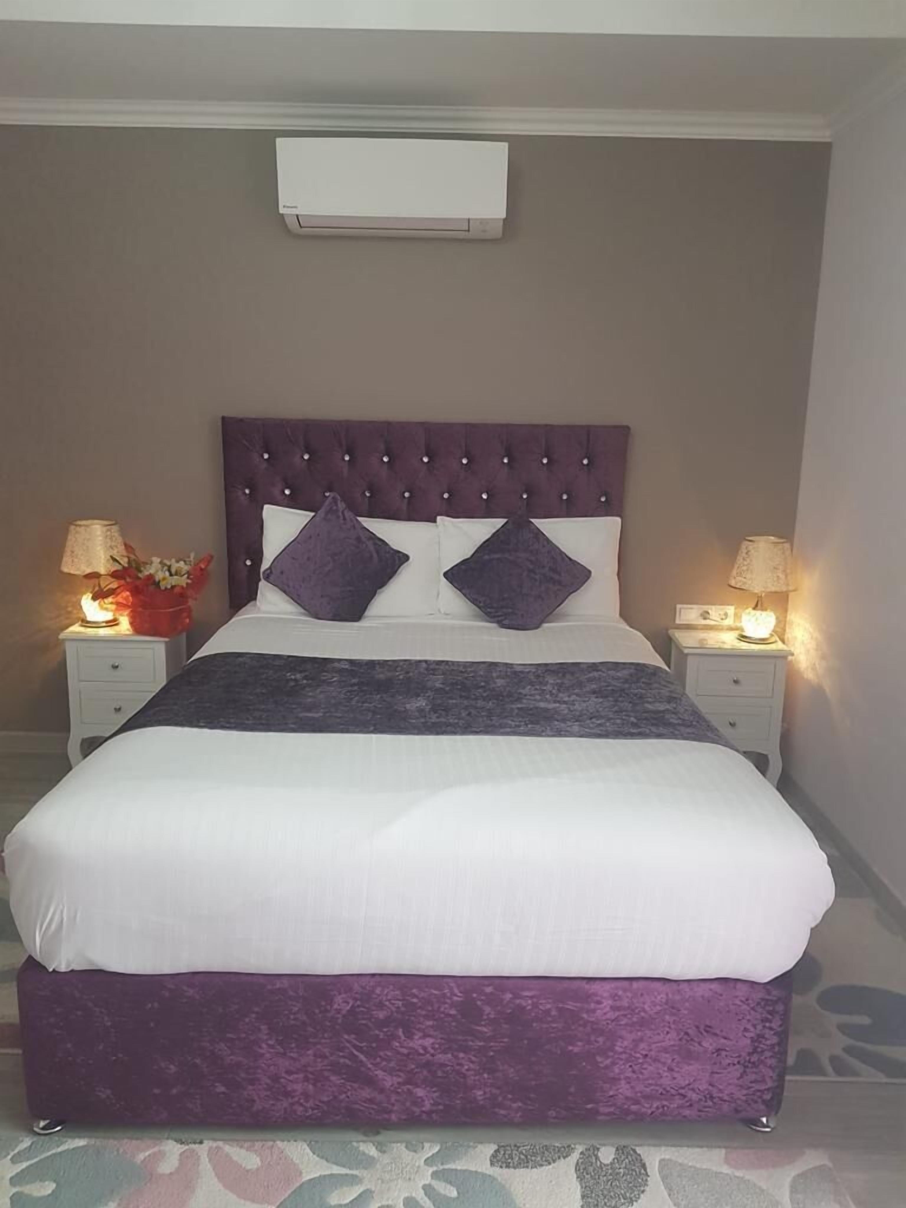 A neatly made bed with a purple headboard, white bedding, and two decorative pillows. There are two bedside tables with lamps and a vase of flowers on each.
