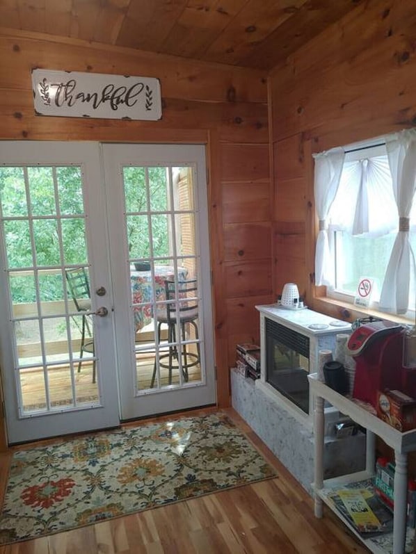 A wooden cabin interior with a sign reading "Thankful" above a door, a small kitchenette, and a view of a deck with chairs.