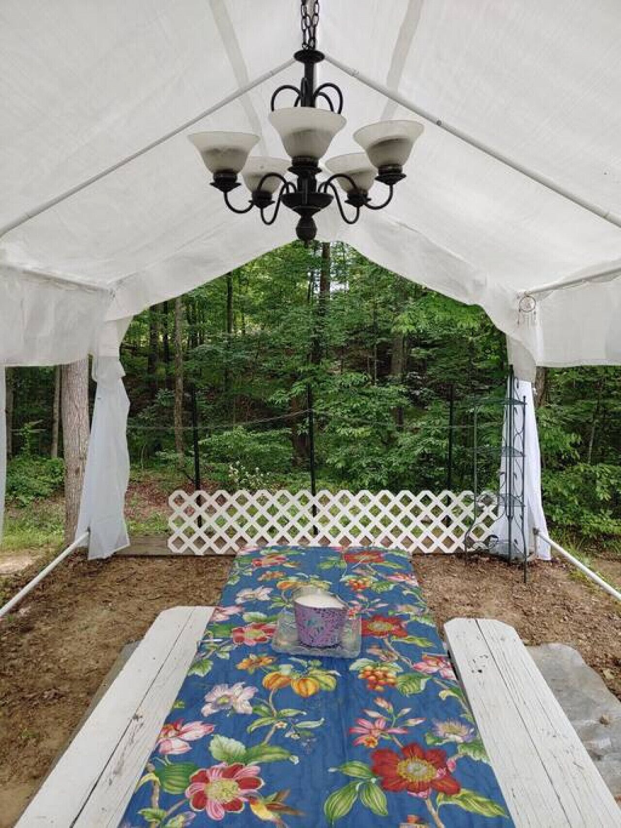 Un espace extérieur abrité, aménagé avec des sièges, une nappe à motifs floraux, une clôture blanche en treillis et un luminaire suspendu.