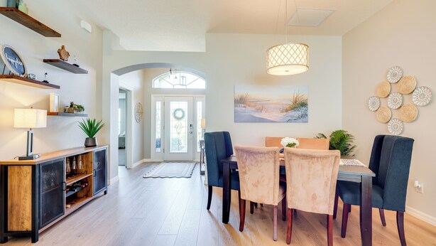 A dining area with a wooden table, chairs, and a pendant light. There are shelves with decorative items and a plant on the left. - Private Lanai & Pool Access: Home in the Villages! (The Villages)