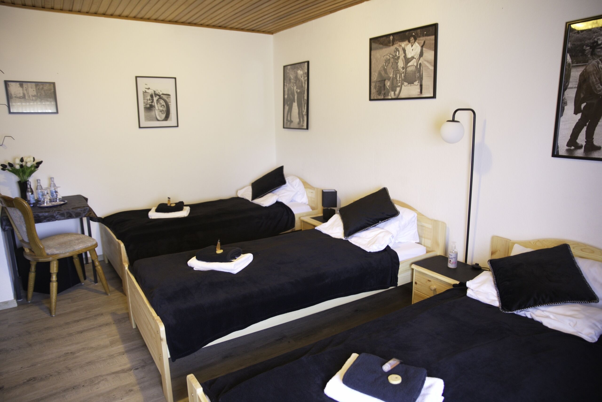A hotel room with three single beds, a desk with a chair, a bedside table with a lamp, and framed pictures on the wall.