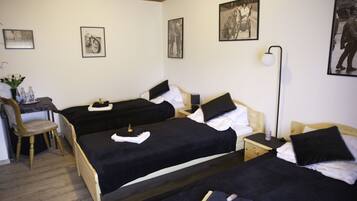 A hotel room with three single beds, a desk with a chair, a bedside table with a lamp, and framed pictures on the wall.