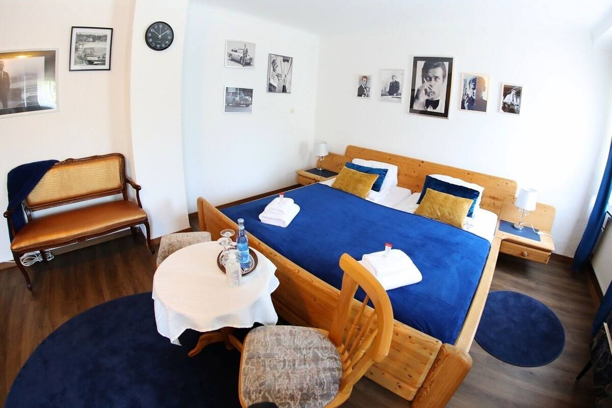A hotel room with a large bed, a wooden chair, a small round table with a bottle and glasses, and a clock on the wall.