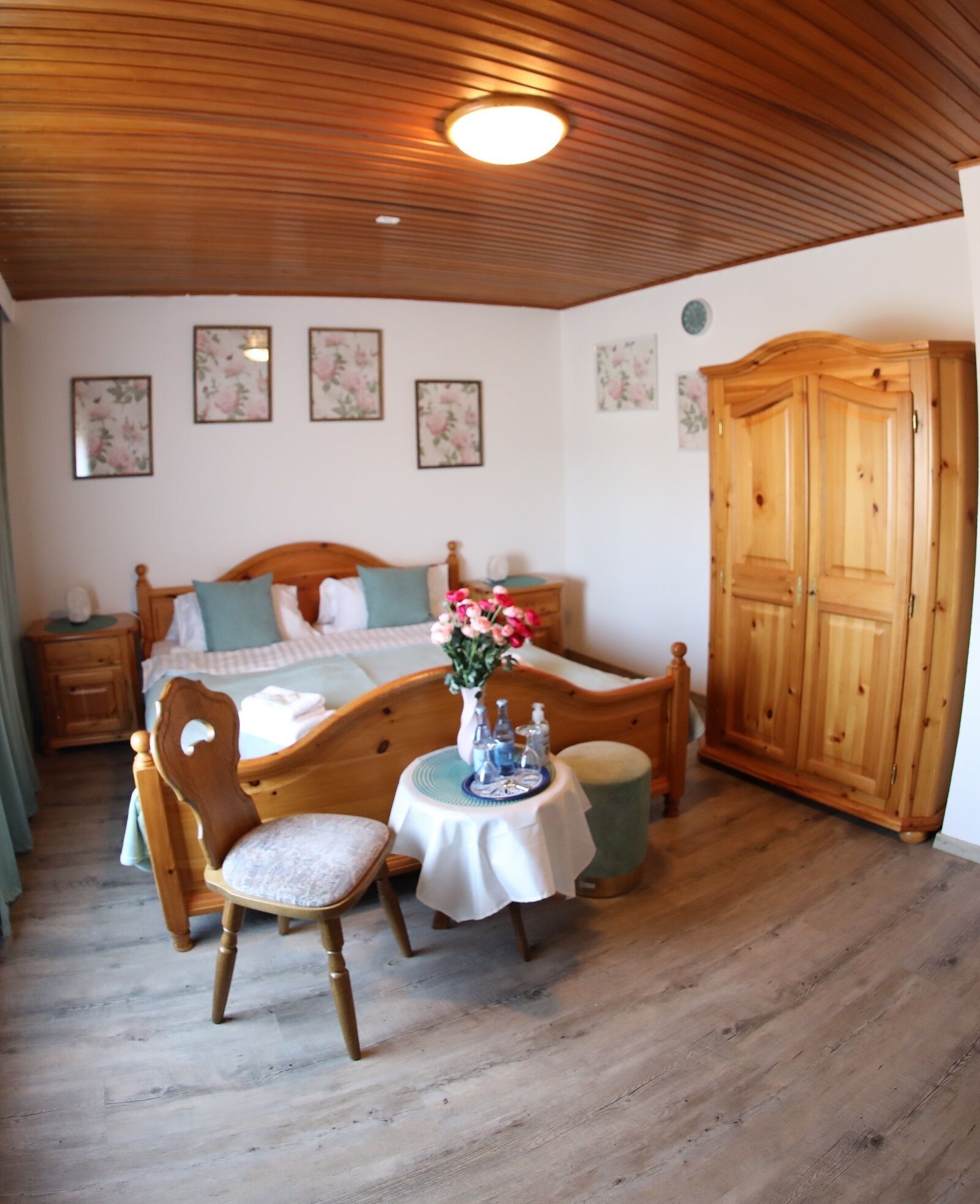 A bedroom with a wooden bed, a bedside table, a chair, a small round table with a vase of flowers, and a wardrobe.