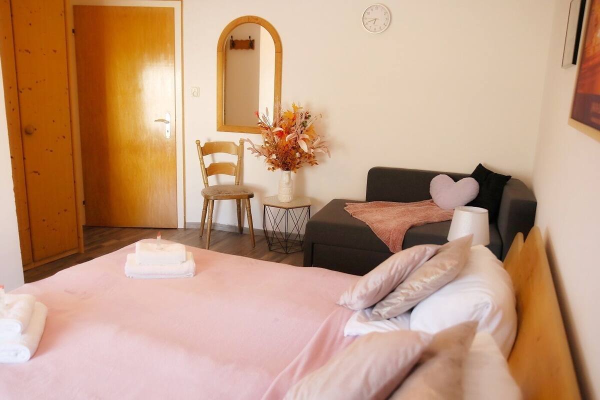 A hotel room with a bed, a chair, a small table with a vase of flowers, and a clock on the wall.