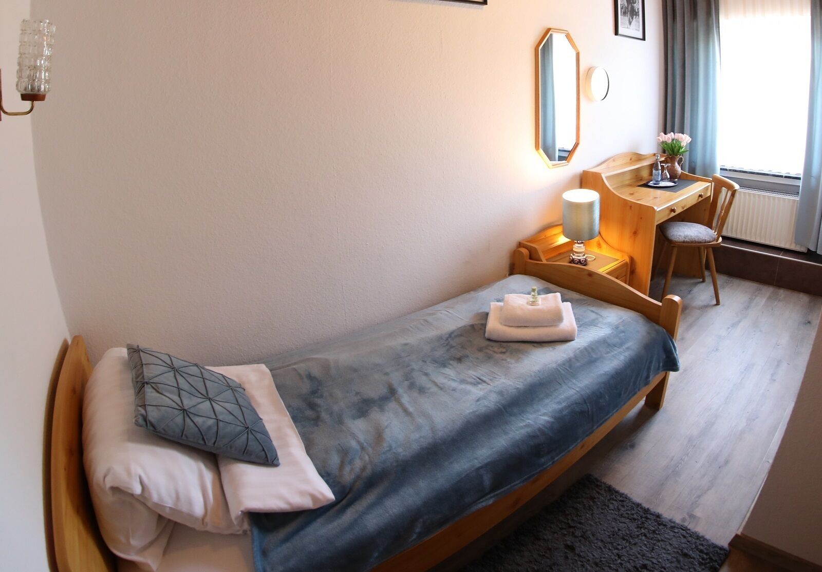 Un dormitorio pequeño y acogedor con una cama individual, un escritorio de madera, una silla y una mesita de noche con lámpara.