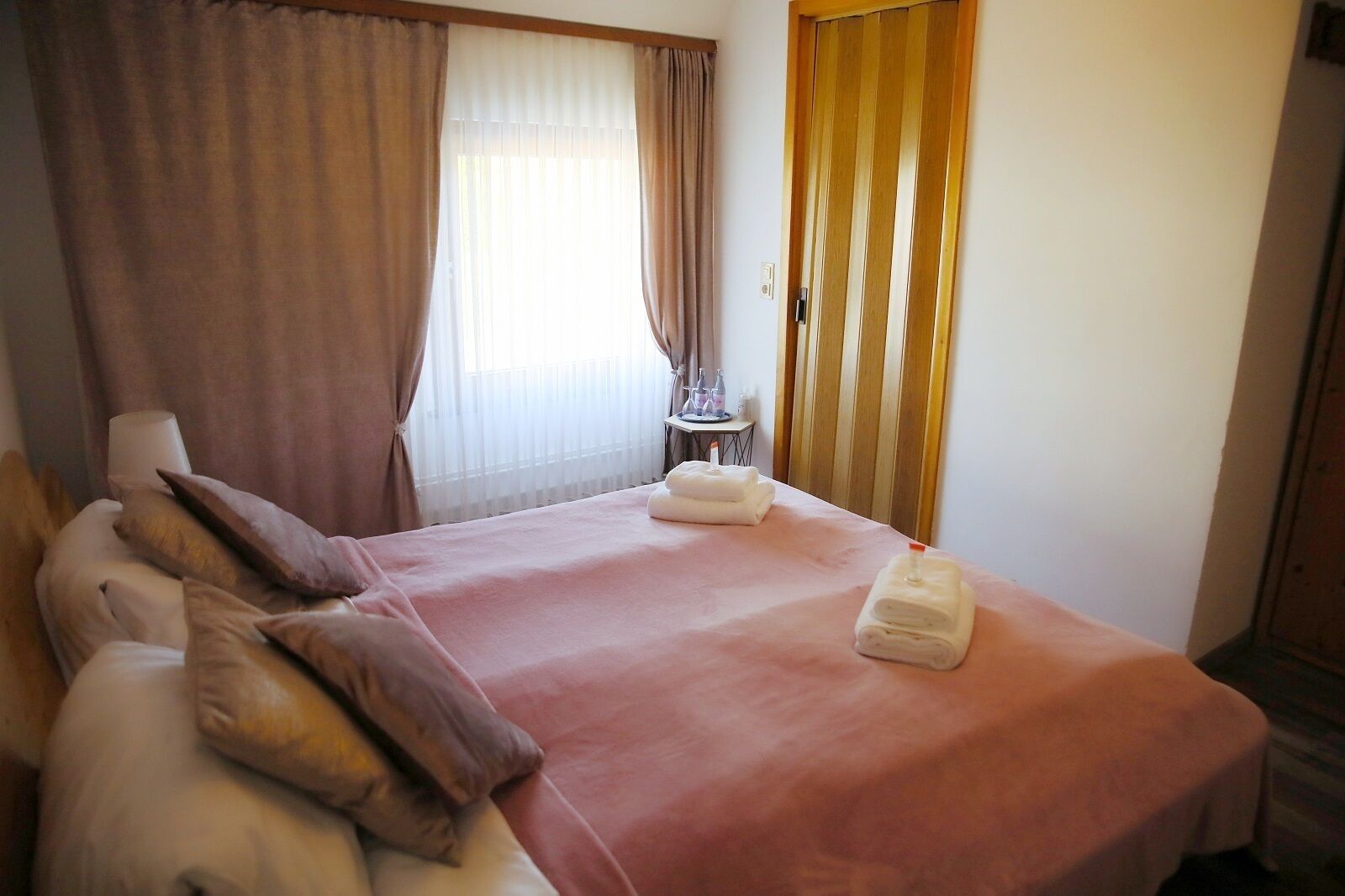 Una habitación de hotel con cama, almohadas, una mesa con botellas y una ventana con cortinas.