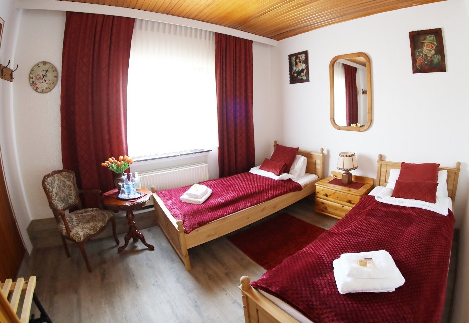 Una habitación con dos camas individuales, una silla de madera, una mesita con un florero y un espejo en la pared.
