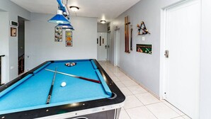 Uma mesa de bilhar com bolas e tacos, um relógio e uma porta em um corredor com luminárias e quadros nas paredes.