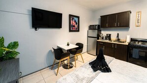 Cozinha compacta com armários escuros, micro-ondas, frigorífico e fogão. Conta com uma pequena área de refeições com mesa e cadeiras. Uma televisão de ecrã plano está fixada na parede acima da área de refeições.