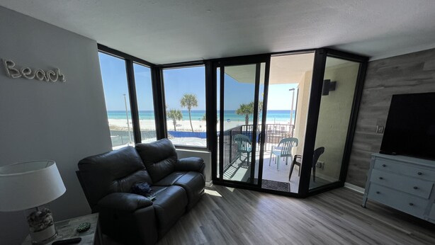A modern living room with a leather sofa, a TV stand, and a view of the beach through large windows. - Complimentary Beach Service with 2 chairs and umbrella (Panama City Beach)
