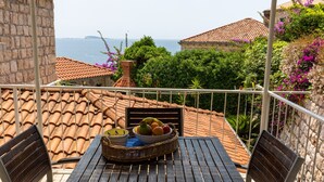 Un balcon avec une table et des chaises, un panier en osier rempli de fruits, et une vue sur la mer et les bâtiments.