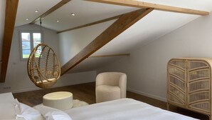 Un dormitorio con techo inclinado, una silla colgante, un sillón mullido, un otoman redondo y una mesita de noche de ratán.