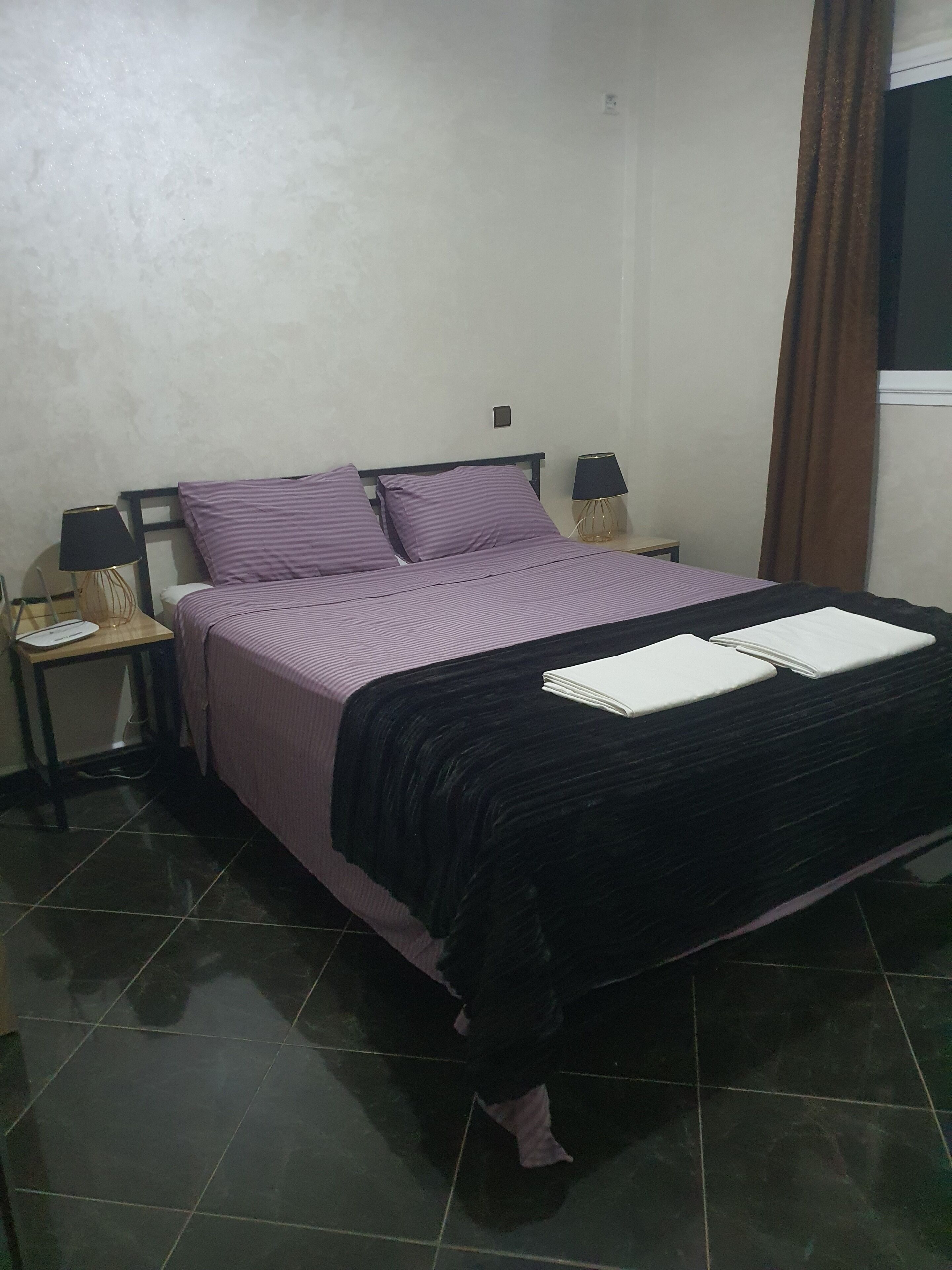 Uma cama com colcha preta e roxa, duas almofadas, uma mesa de cabeceira com abajur e uma janela com cortinas marrons.