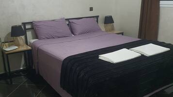 Uma cama com colcha preta e roxa, duas almofadas, uma mesa de cabeceira com abajur e uma janela com cortinas marrons.