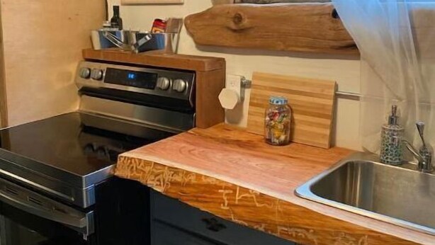 A compact kitchen with a stove, oven, and sink. A wooden countertop extends from the sink, and a window with a sheer curtain is above the sink. - A Cabin Crafted for Retreat, Celebration & Everyday Getaways (Powell River)