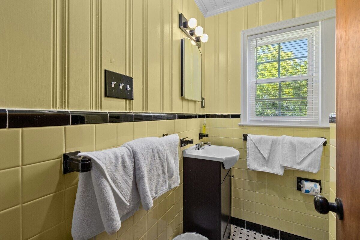 Ein Badezimmer mit gelben Wänden, einer schwarzen Duschkabine und einem weißen Waschbecken, sowie einem Fenster mit Jalousien.