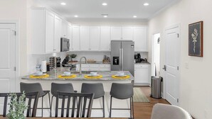 A modern kitchen with white cabinets, a central island with bar stools, stainless steel appliances, and recessed ceiling lighting. - Landing Zone | Pet-Friendly 4BR w/ Pool + Hot Tub (North Myrtle Beach)