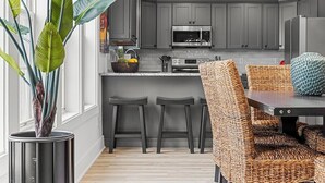 A modern kitchen with grey cabinets, a microwave, and a dining area with wicker chairs. - Hem's Hideout | 5BR Tropical Oasis w/ Heated Pool (North Myrtle Beach)