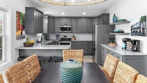 A modern kitchen with grey cabinets, a central island, and a dining area with wicker chairs. - Hem's Hideout | 5BR Tropical Oasis w/ Heated Pool (North Myrtle Beach)
