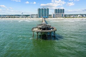 Un muelle se adentra en el agua con un pabellón en el centro, rodeado por una playa y edificios de gran altura al fondo.