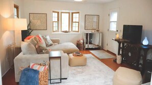 A living room with a sofa, ottoman, and a TV stand. There are windows, a lamp, and a decorative mirror.