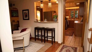 A cozy living room with a white sofa, two dark barstools, a coffee table, and a fireplace. A wooden door leads to a kitchen with wooden cabinets and a tiled floor.