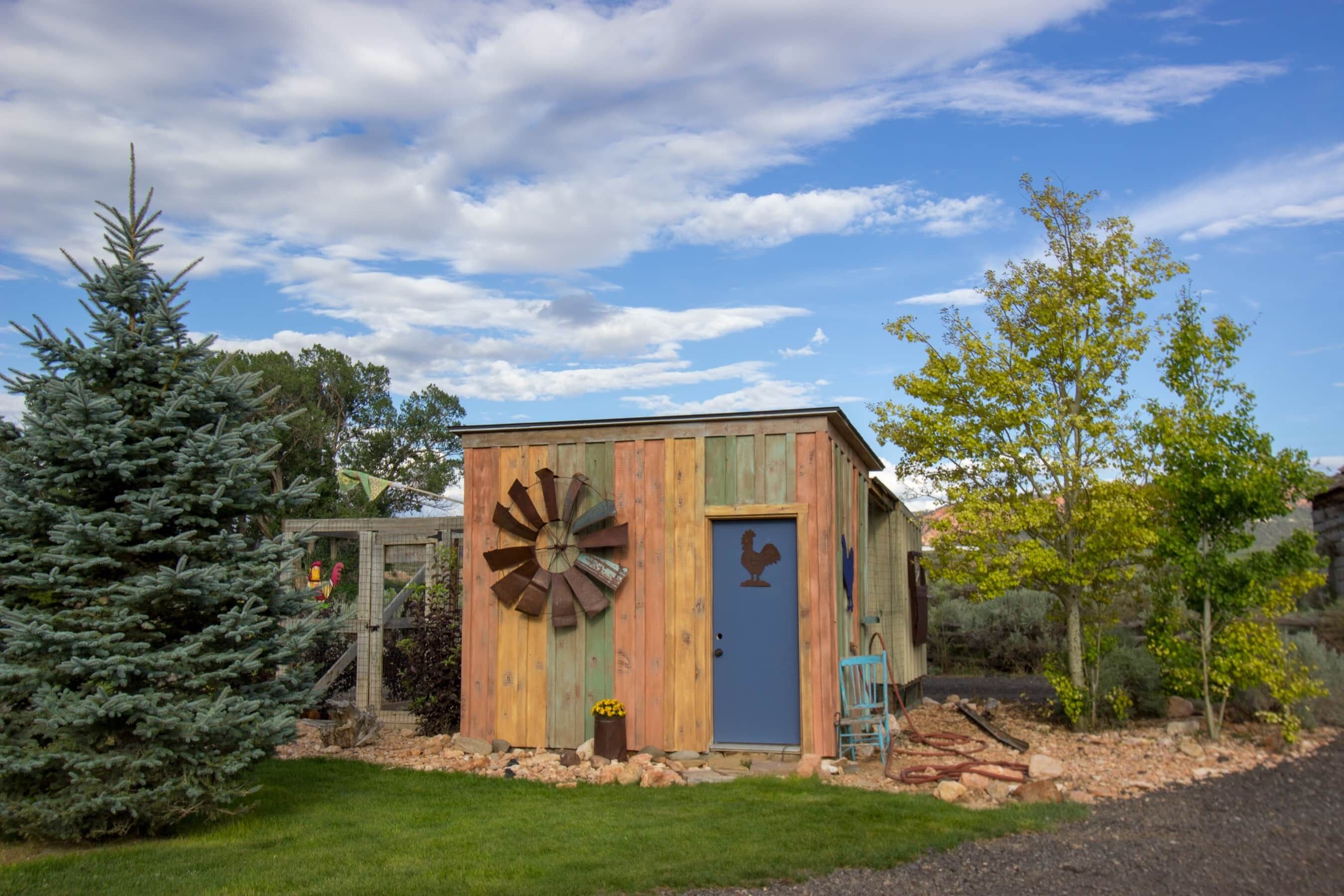 A rustic wooden building with a windmill, a rooster door, and a small garden area.