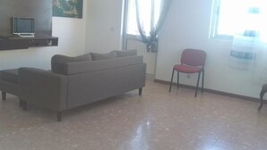 A living room with a grey sofa, a red chair, and a brown chair. There is a TV on a shelf and a window with curtains. - Cubico Tota (Praia)