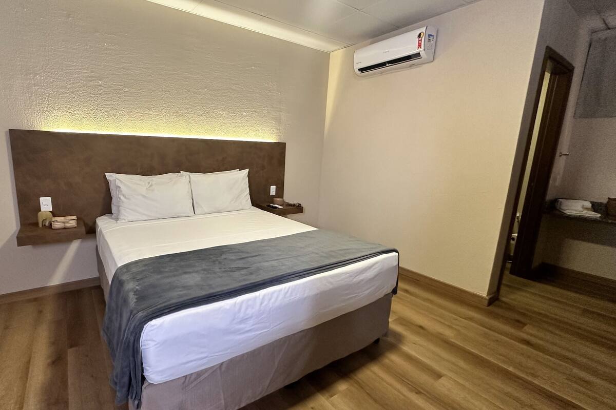 A hotel room with a bed, bedside tables, an air conditioning unit, and a wooden floor.