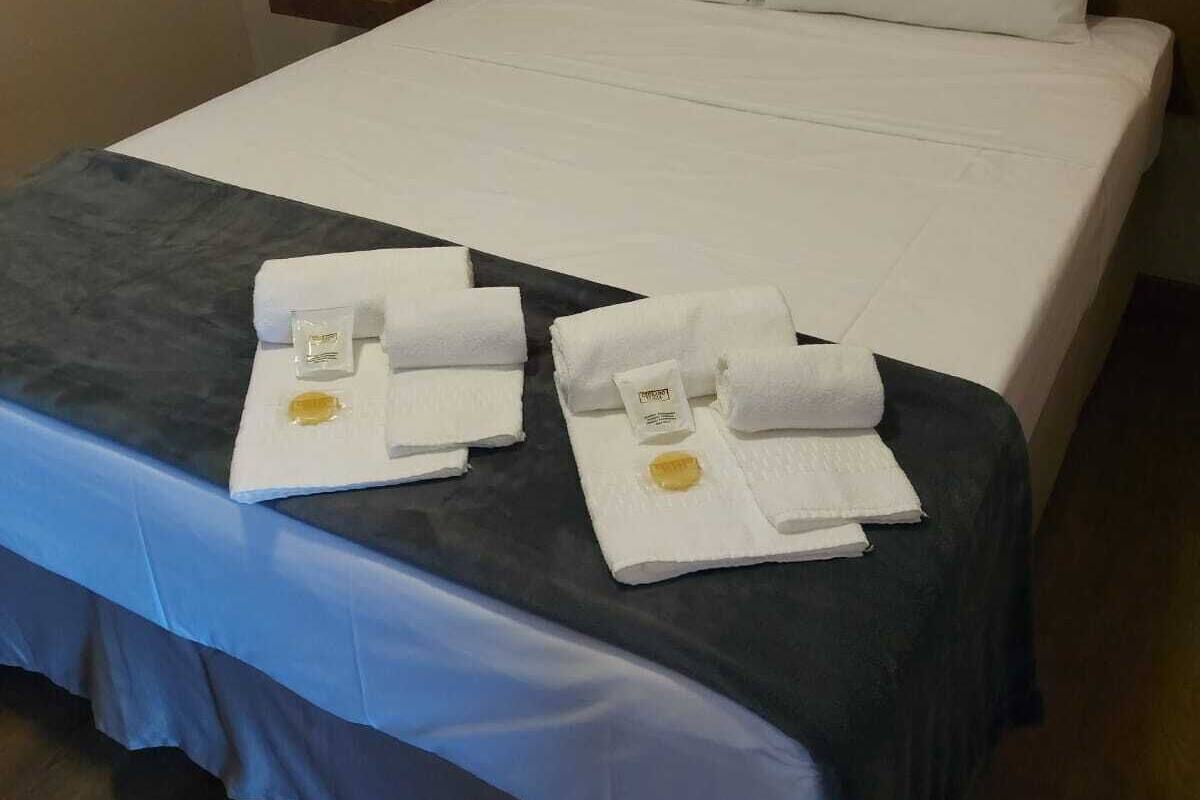 A neatly made bed with white linens, a blue bed skirt, and two towels with a yellow emblem on a wooden nightstand.