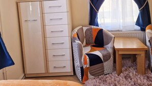 Ein Zimmer mit einem weißen Kleiderschrank, einem an der Wand befestigten Fernseher, einem gemusterten Sessel, einem kleinen Holztisch und einem Fenster mit blauen Vorhängen.