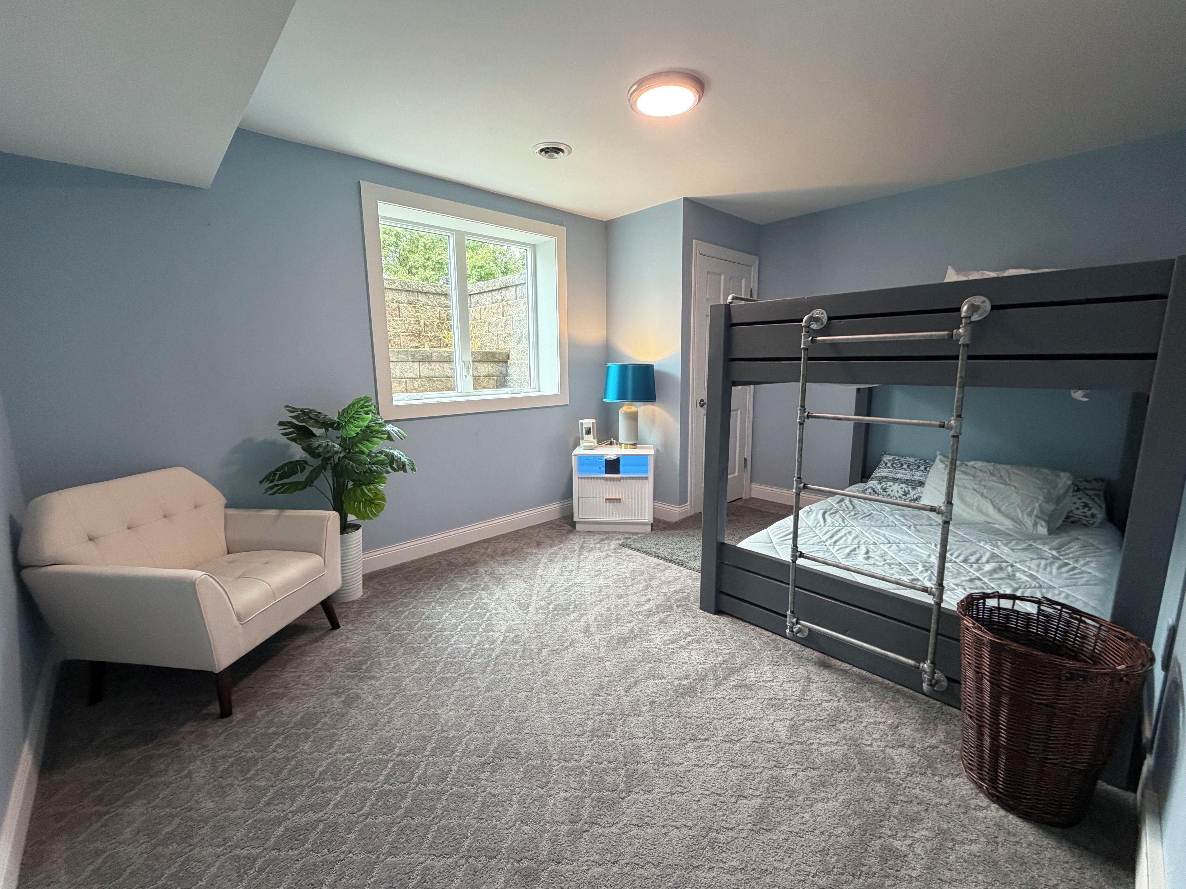 A bedroom with a bunk bed, a chair, a bedside table with a lamp, and a potted plant.