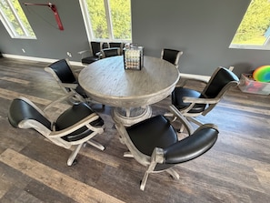 A round dining table with six chairs, a decorative centerpiece, and a window with a view of trees.