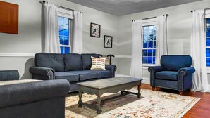 A living room with a dark blue sofa, a matching armchair, a coffee table, and a patterned rug. There are two windows with white curtains and two framed pictures on the wall. - Rivers Edge (Freedom)