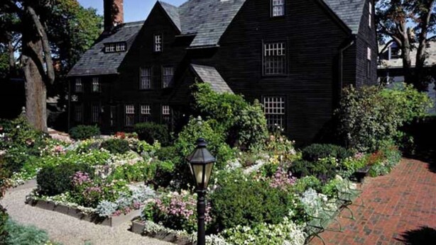 A traditional black wooden house with multiple gabled roofs, surrounded by a well-maintained garden with various flowering plants and a brick pathway.