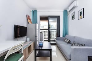 A compact living space with a kitchenette, a grey sofa, a flat-screen TV, and a balcony with a view.