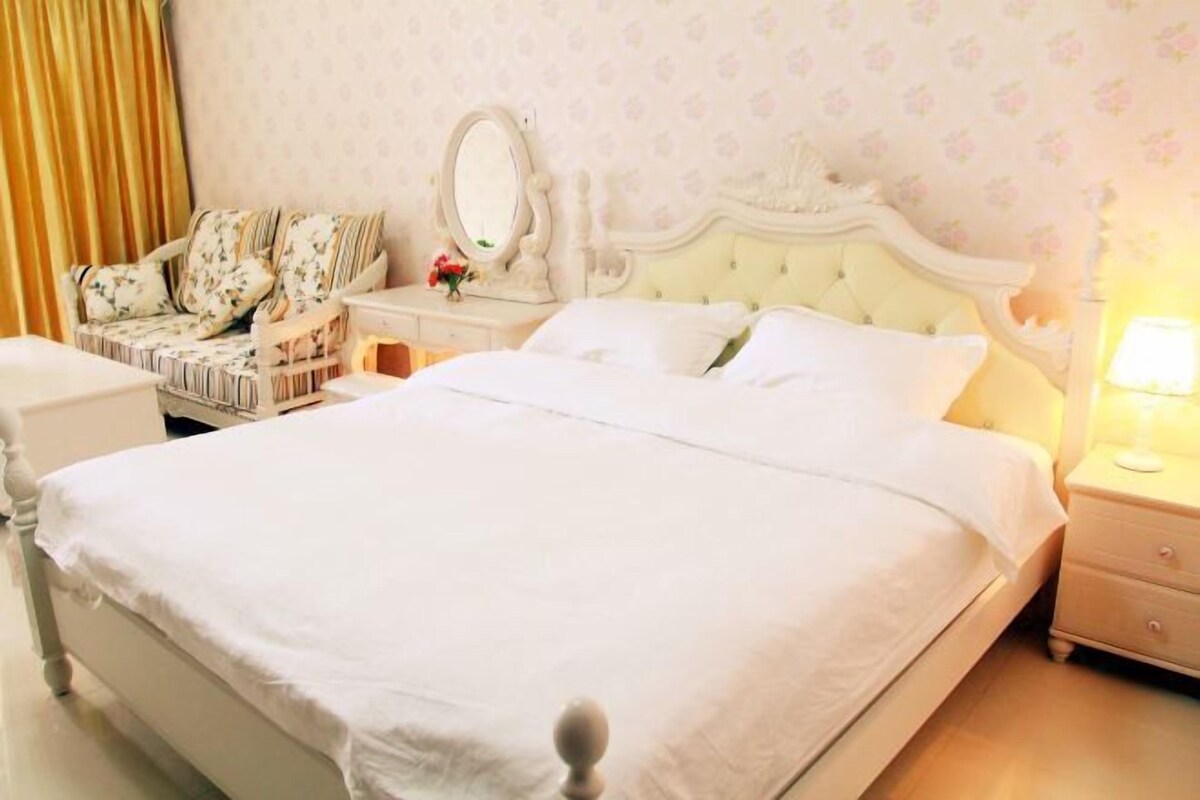 A neatly made bed with a white headboard, a bedside table with a lamp, and a floral-patterned armchair.