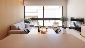 A modern living room with a sofa, a dining table with a bottle and fruit, a TV, and large windows. - Bravissimo Centre, Modern 2-bedroom apartment (Girona)