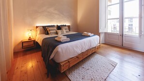 A bedroom with a wooden bed, a nightstand with a lamp, a large window, and a view of buildings outside. - Bravissimo Carrer Nou, cosy 2-bedroom apartment in the center of Girona (Girona)