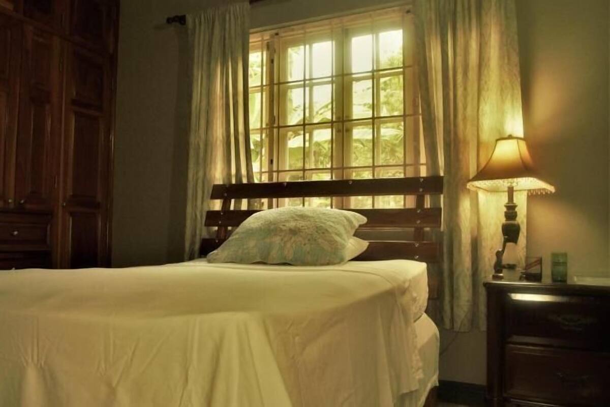 A bedroom with a wooden bed, a nightstand with a lamp, and a window with curtains.
