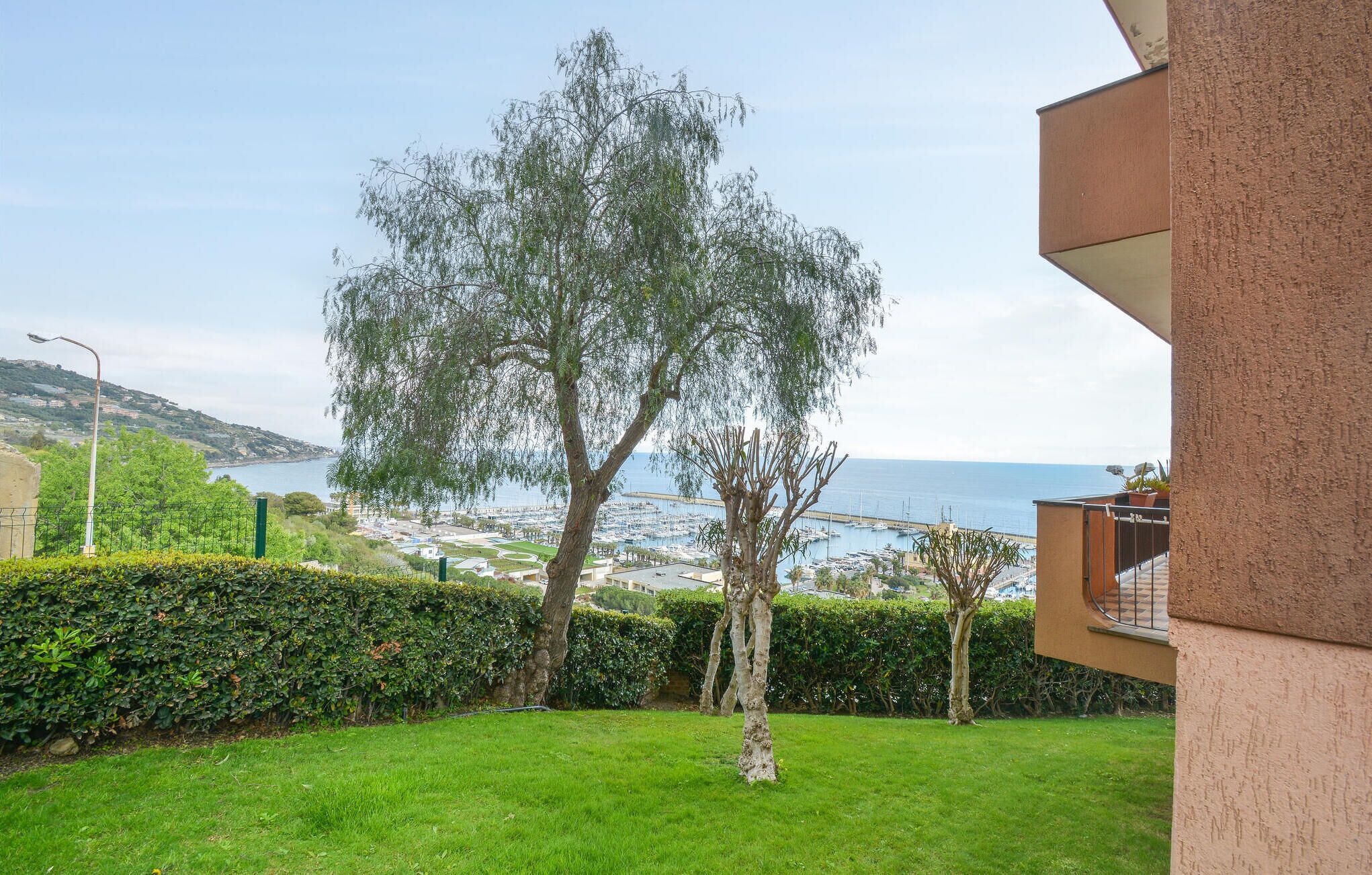 A view of a coastal area with a large tree in the foreground, a grassy lawn, and a building with a balcony.