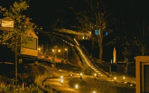 A nighttime outdoor area with a pathway leading up a hill, flanked by trees and illuminated by street lamps. - Bobocabin Gunung Mas (Cisarua)