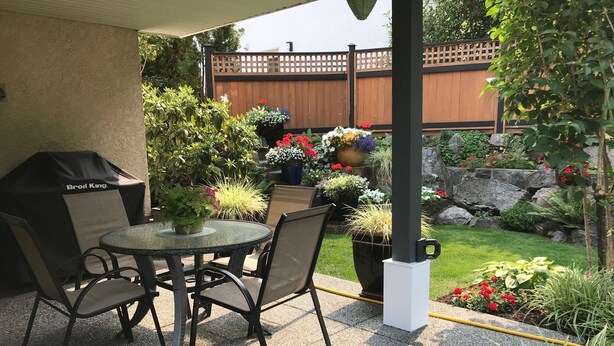 En uteplats med ett runt bord och stolar, en grill samt olika krukväxter och blommor.