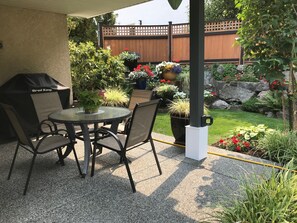Un patio con una mesa redonda y sillas, una parrilla y varias macetas con plantas y flores.