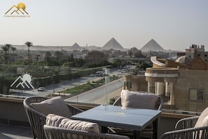 A balcony with wicker chairs and a table, overlooking a road with cars and a view of the pyramids in the distance.