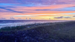 Een kustlandschap met een zonsondergang boven de oceaan, grasduinen en een pier in de verte.